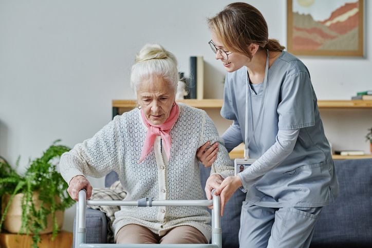 Caregiver assists a woman using a walker to stand; living room setting.