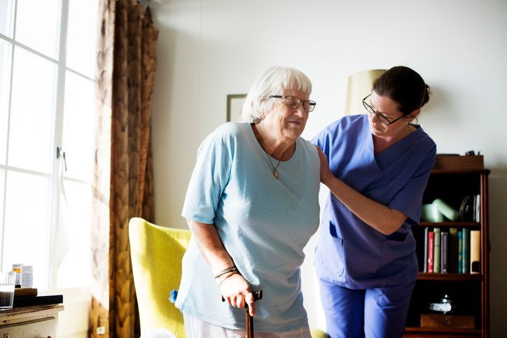Caregiver assisting an elderly person with a cane to stand in a well-lit living room.