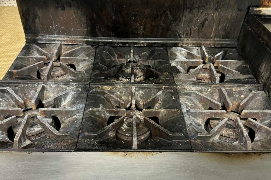 Heavy carbonized grease and burnt-on residue covering the burner grates and burner heads of a six-burner commercial kitchen range before cleaning