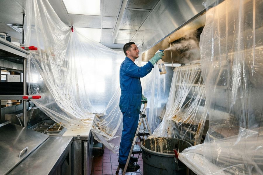 Technician on a step ladder spraying degreaser into a commercial kitchen exhaust hood, with plastic sheeting draped over the equipment below and a waste barrel catching runoff