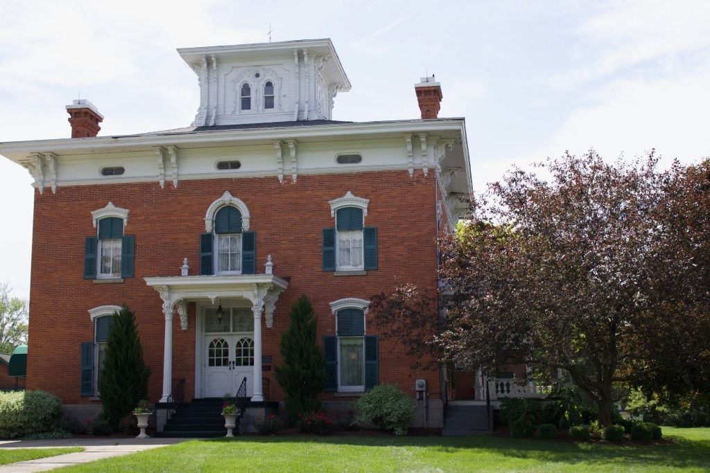 Red brick house with white trim, green shutters, and a cupola. Lush green lawn and tree in front.