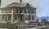 Two-story house with shutters, porch, and a horse standing nearby. A fence and garden in the foreground.