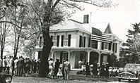 Black and white photo of a large white house with many people gathered in front.