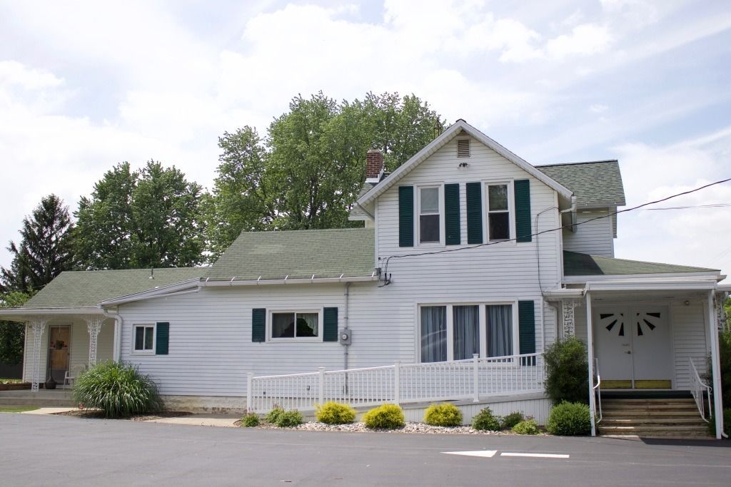 White two-story building with green shutters, a porch, and a ramp. Trees and a paved area are in front.