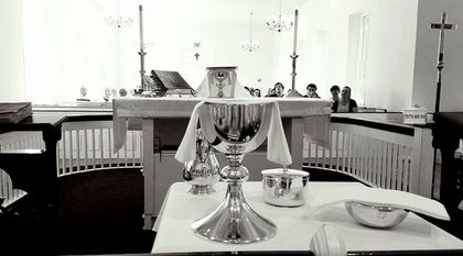 A silver chalice sits on a small table in front of a church altar, with a small group of people seated in the background.