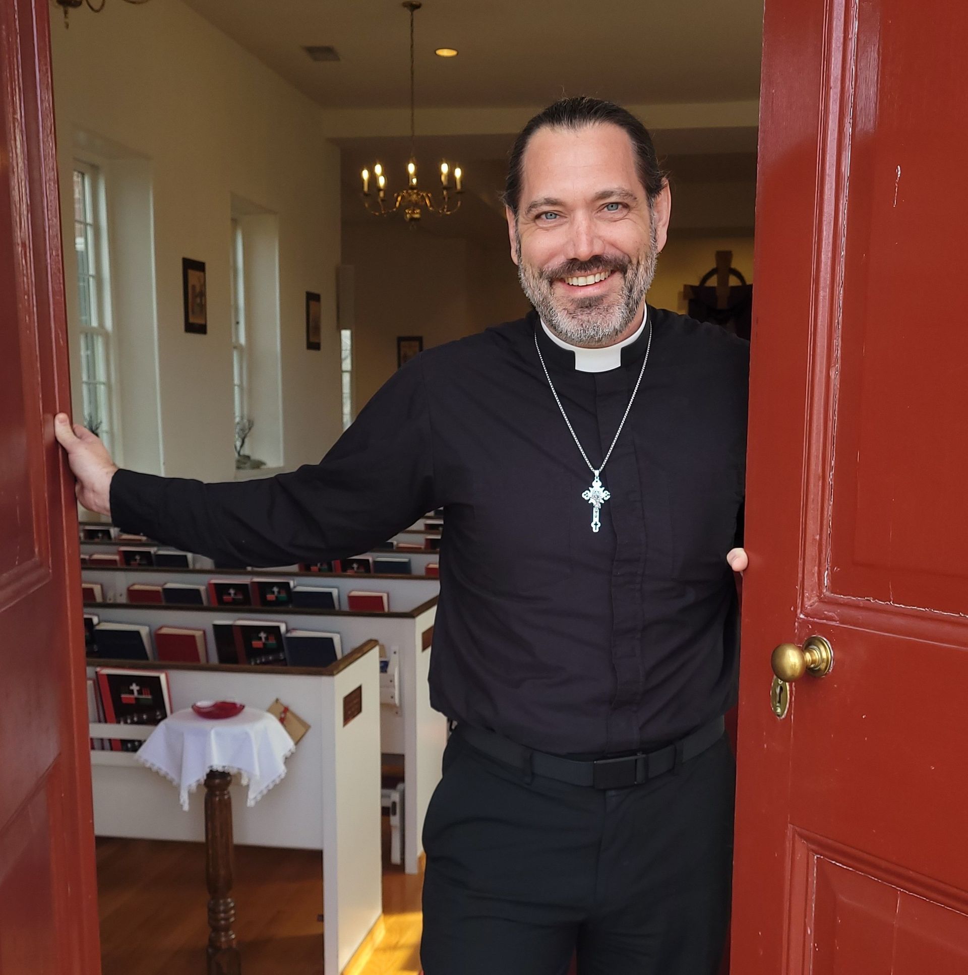 A smiling priest in a black shirt and clerical collar stands in an open red doorway of a church with rows of pews.