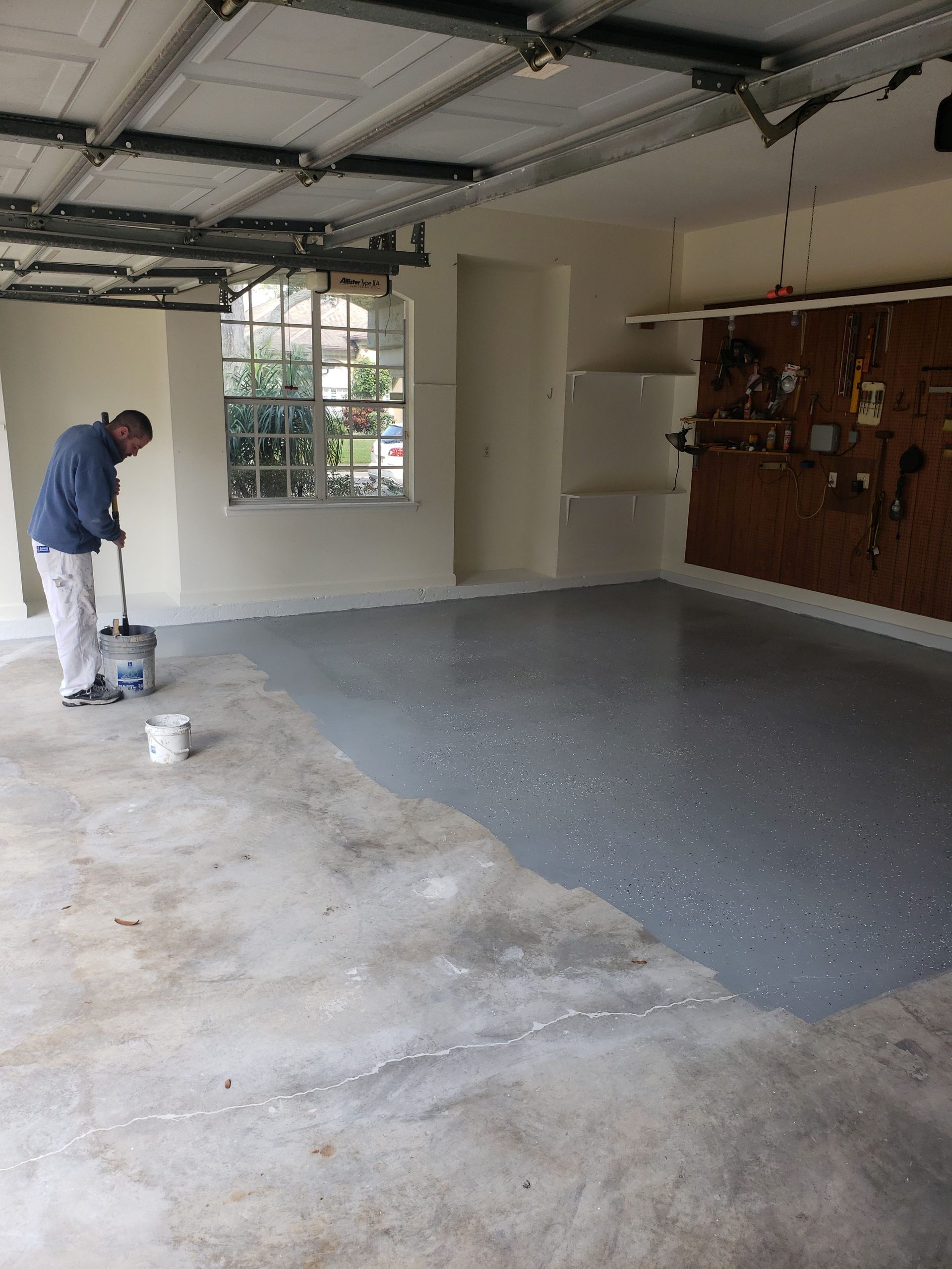 Person painting a garage floor gray, with tools and an open garage door visible.