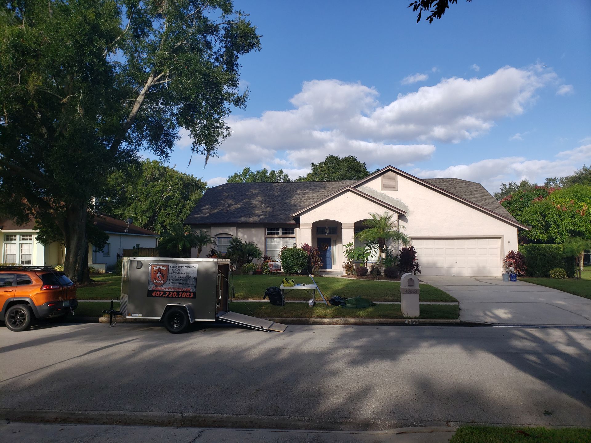 Suburban house with white stucco exterior, parked trailer and orange truck, and a driveway on a sunny day