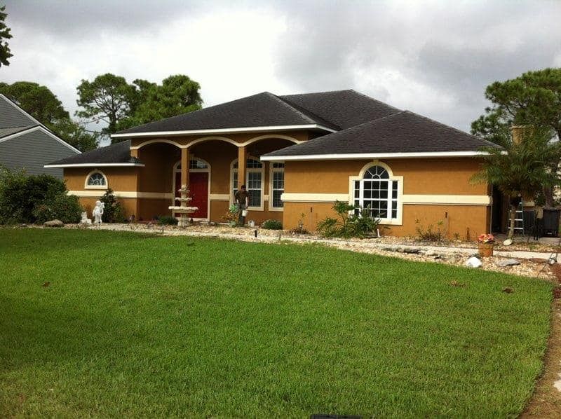 Single-story brick house with a dark roof and large green lawn under cloudy skies