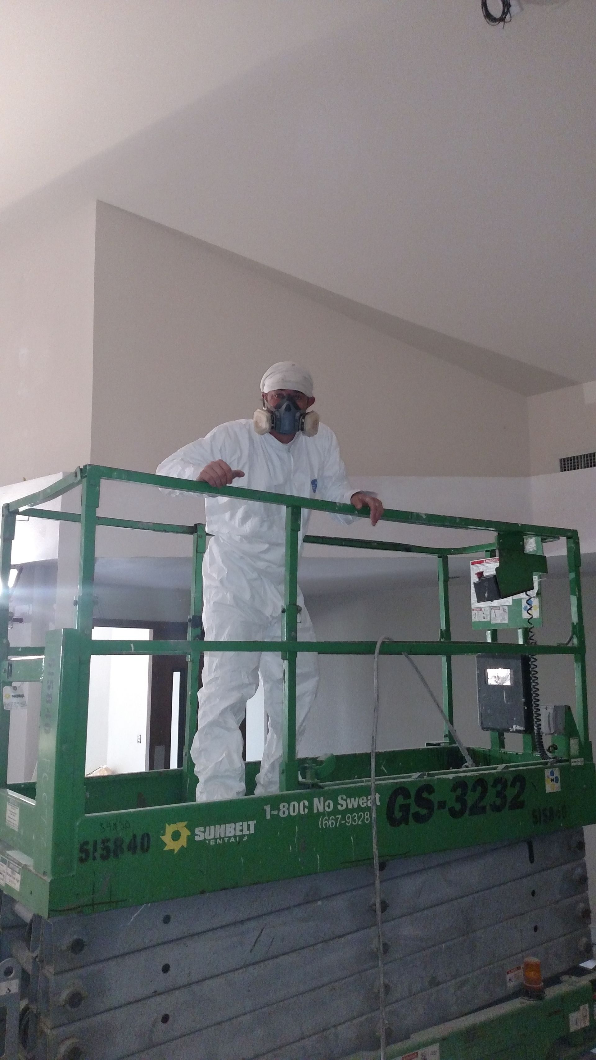 Worker in white protective suit standing on a green lift platform indoors, cleaning or painting a wall.