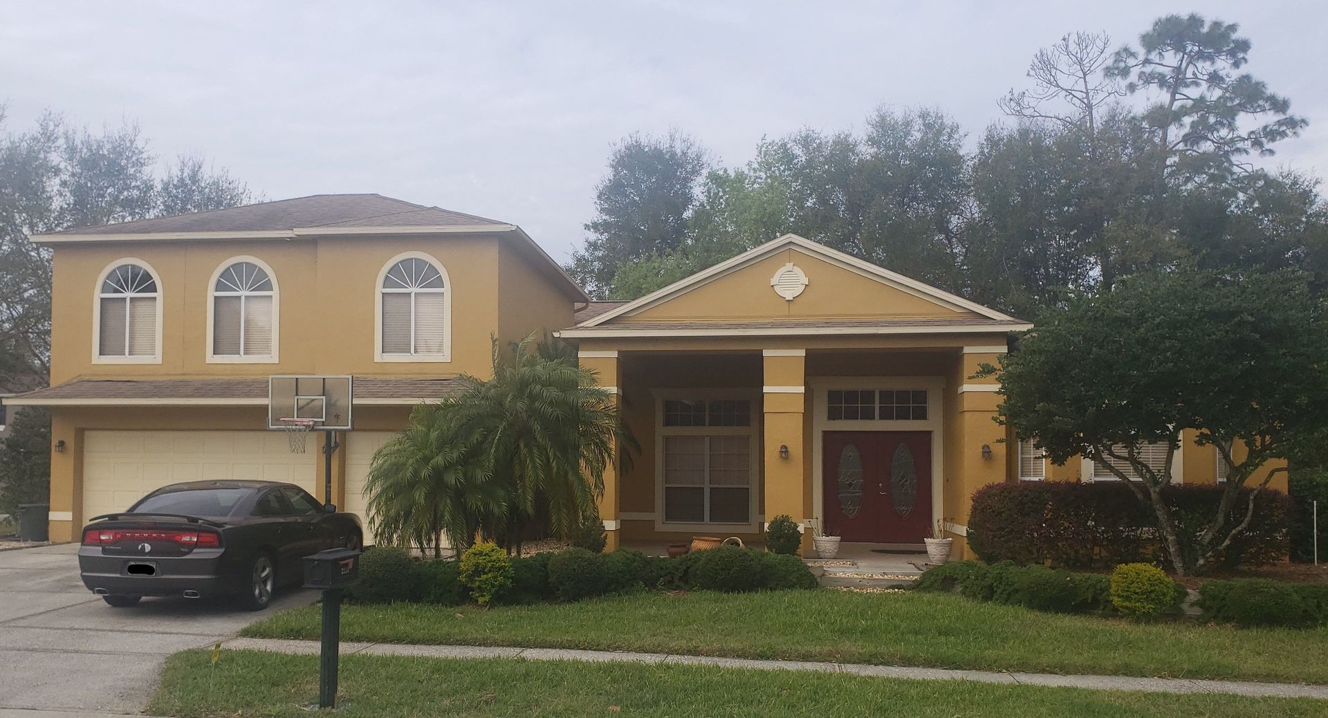 Yellow two-story house with a red front door, dark car in driveway, and overcast sky