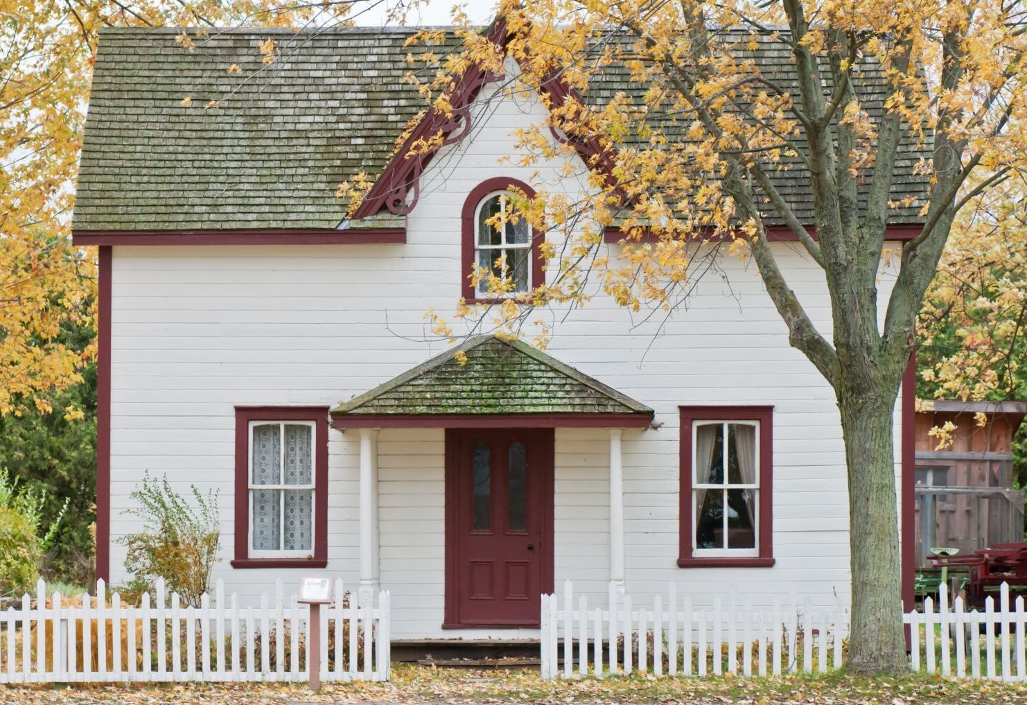 Small white house with red door, porch, and picket fence under autumn trees