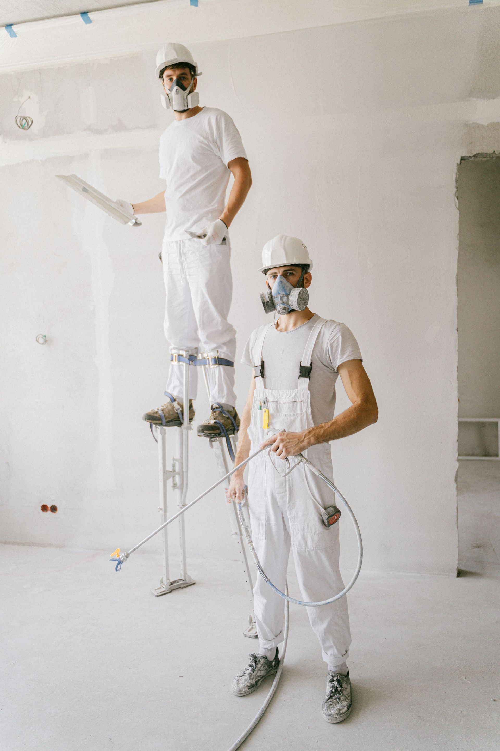 Two painters in white work clothes on metal scaffolds, painting a white interior wall.