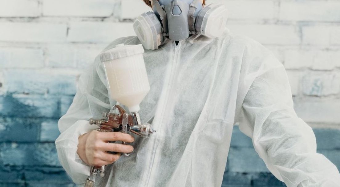 Person in a white protective suit spraying paint with a spray gun against a brick wall.