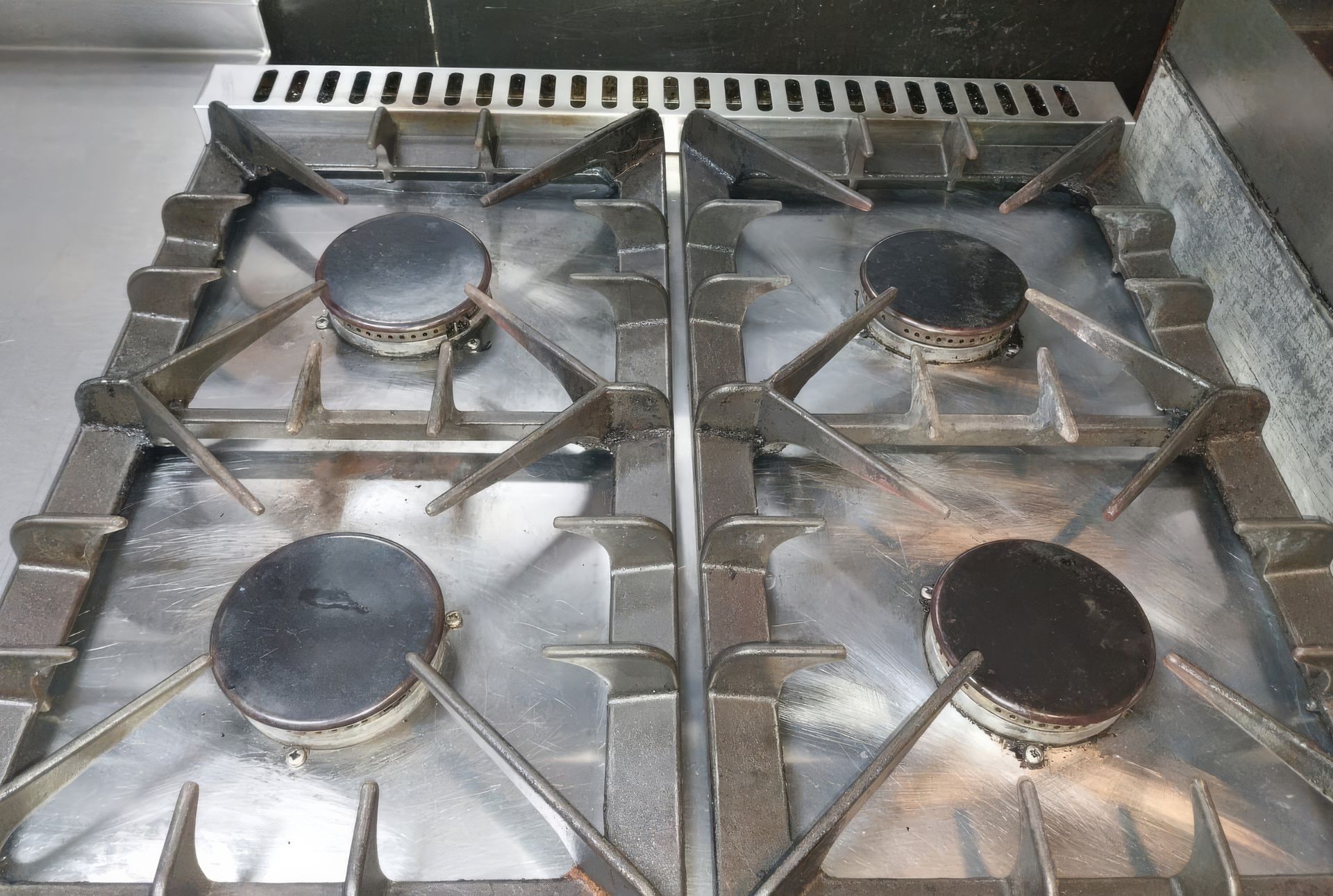 Stainless steel commercial gas stovetop with four burners, looking slightly dirty.
