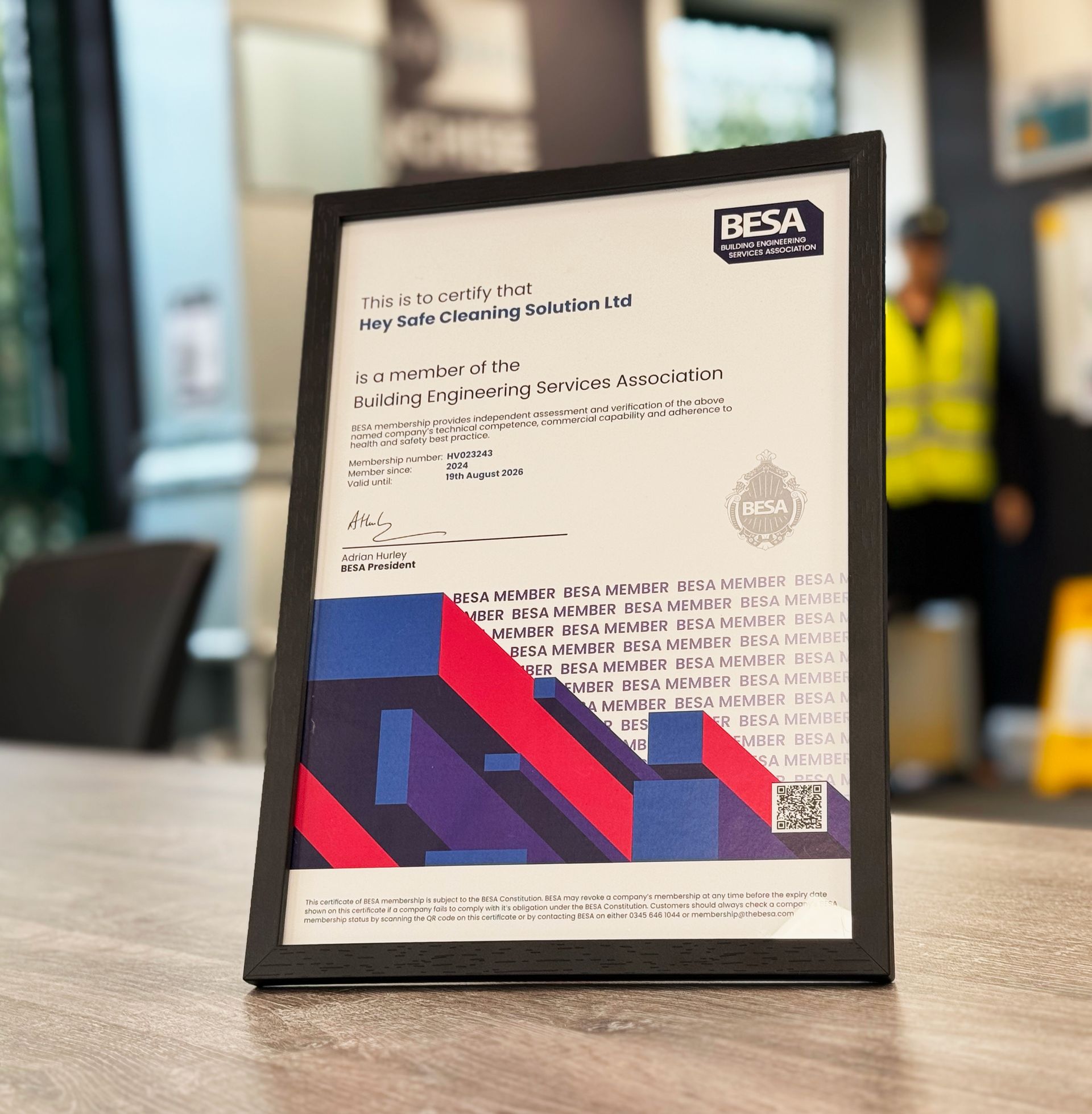 Black framed certificate with BESA logo on a table. Person in background wearing a high-vis vest.