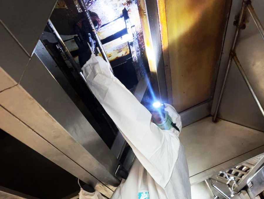 Worker in white protective suit inspecting machinery inside a narrow industrial space, lit by a flashlight.