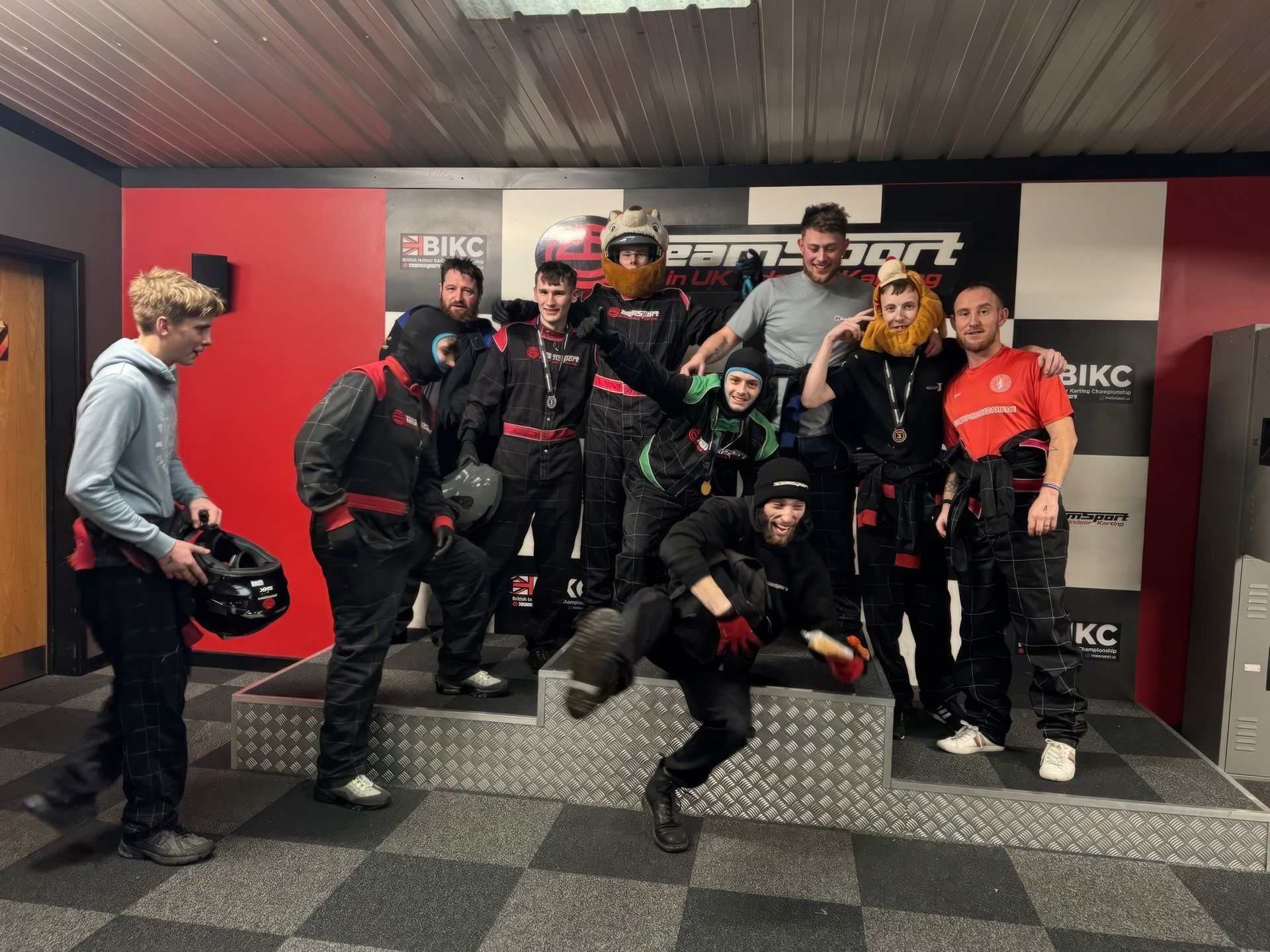 Group of people in racing suits posing on a podium at a go-kart track, some smiling.