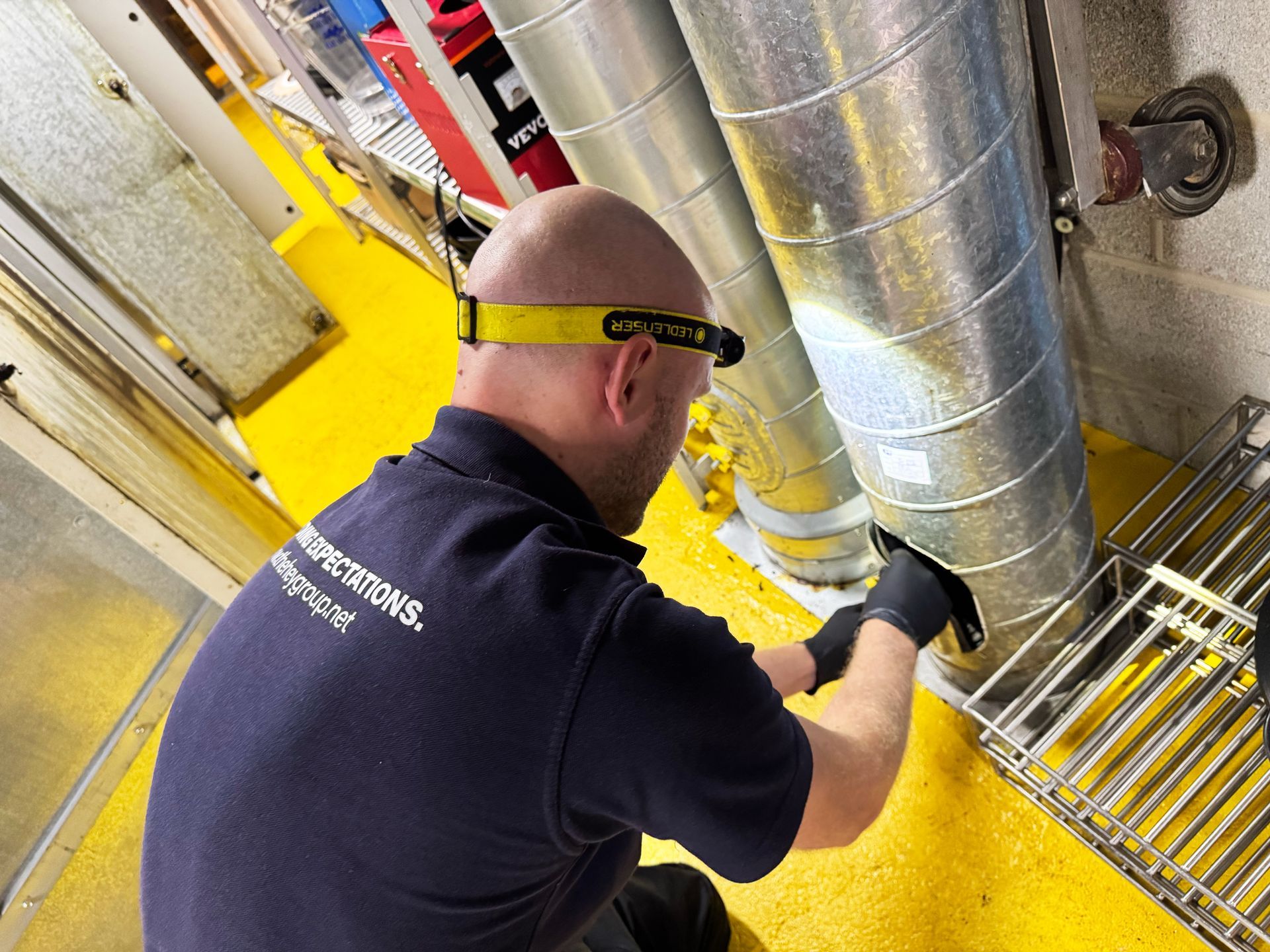 Man in a dark blue shirt and gloves inspects ductwork with a headlamp in a yellow-floored industrial setting.