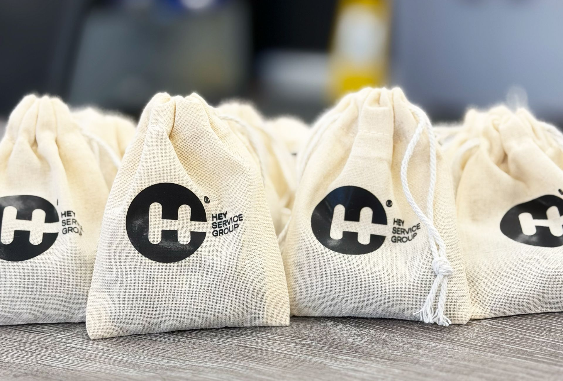 Cream-colored drawstring bags with a black logo, grouped on a surface. The logo is a stylized