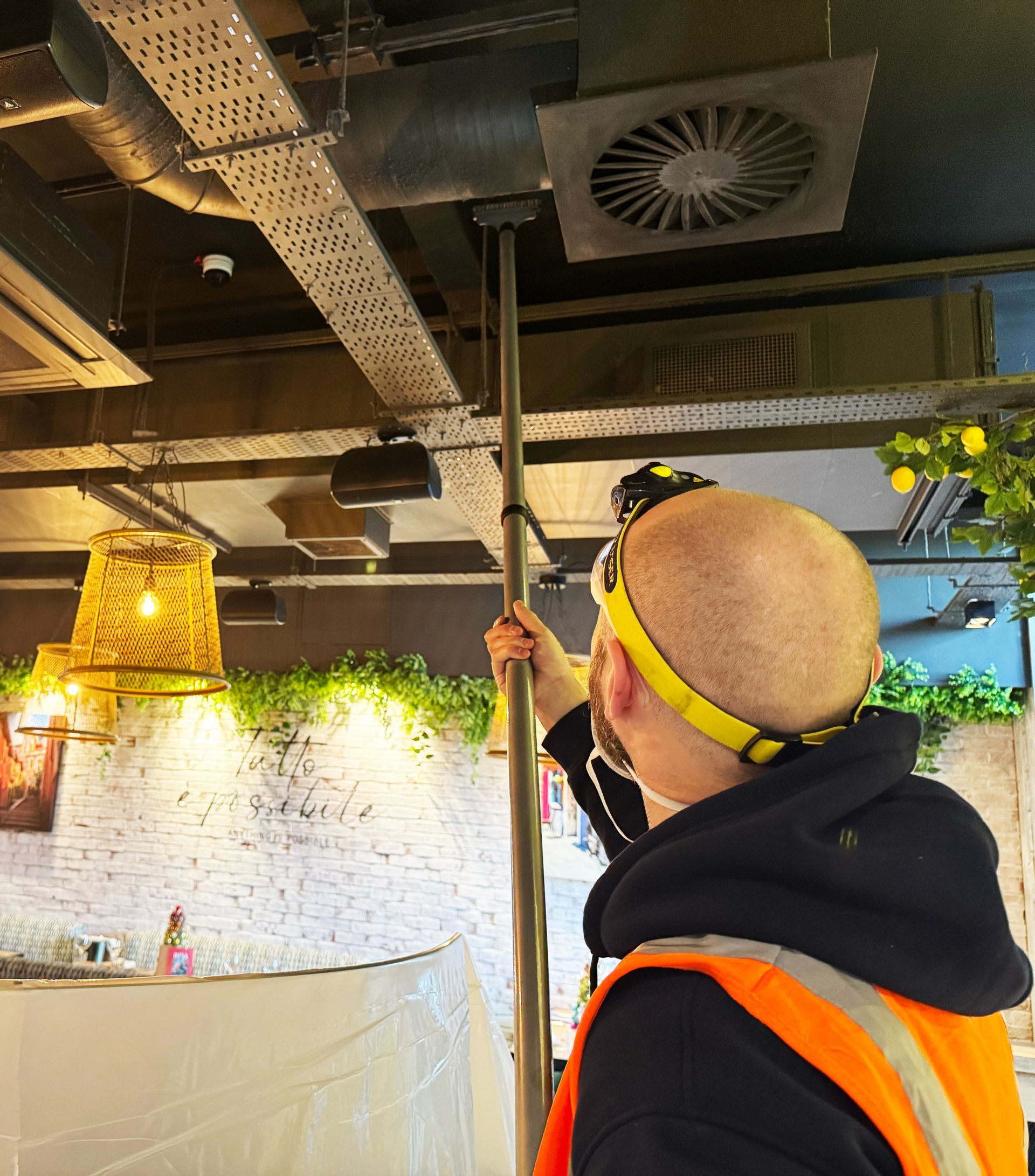 Worker in an orange safety vest using a long pole under an industrial ceiling with vents and hanging lights