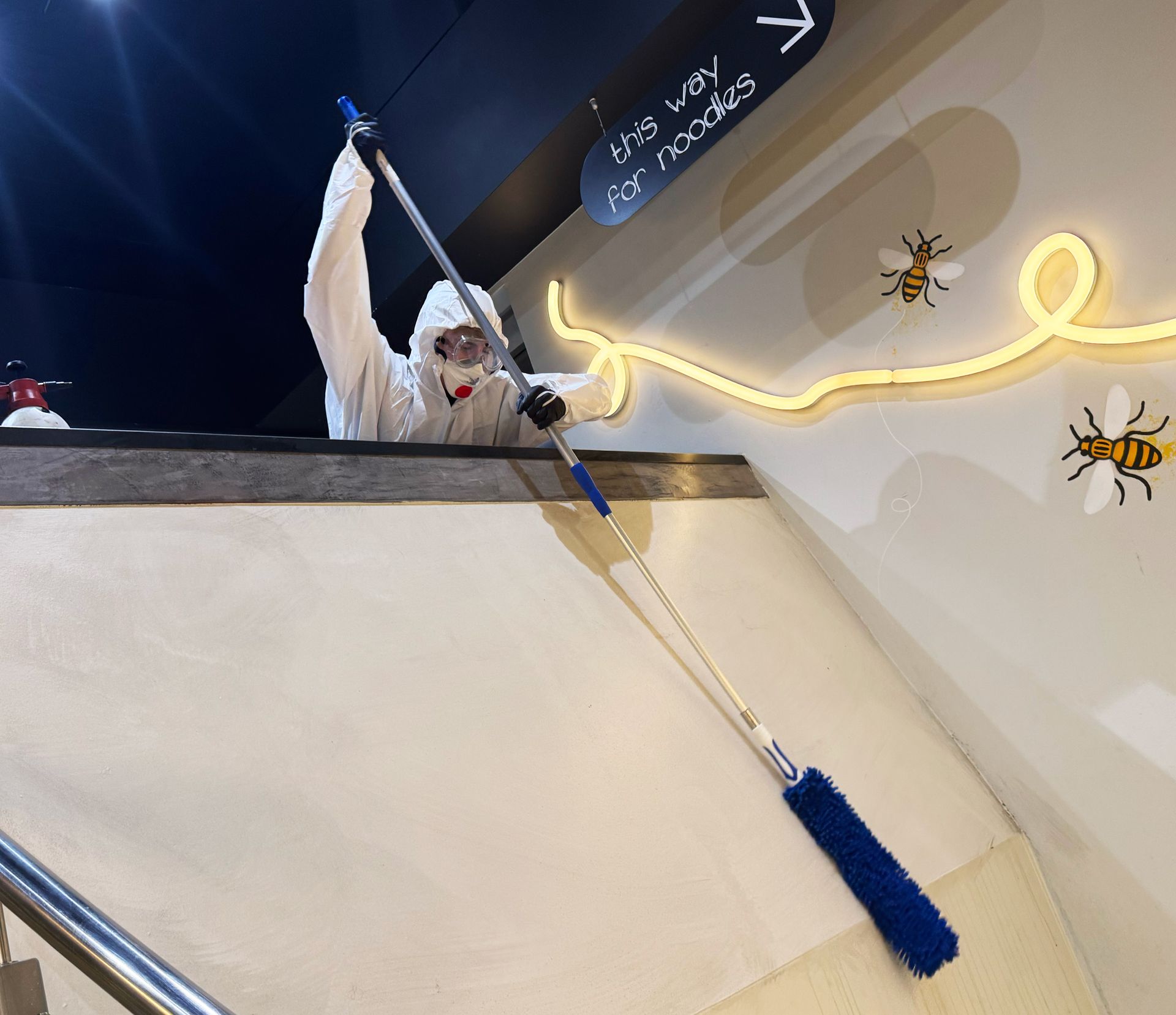 Person in protective suit cleaning a stairwell with a long-handled duster; a sign and decorative bees are nearby.