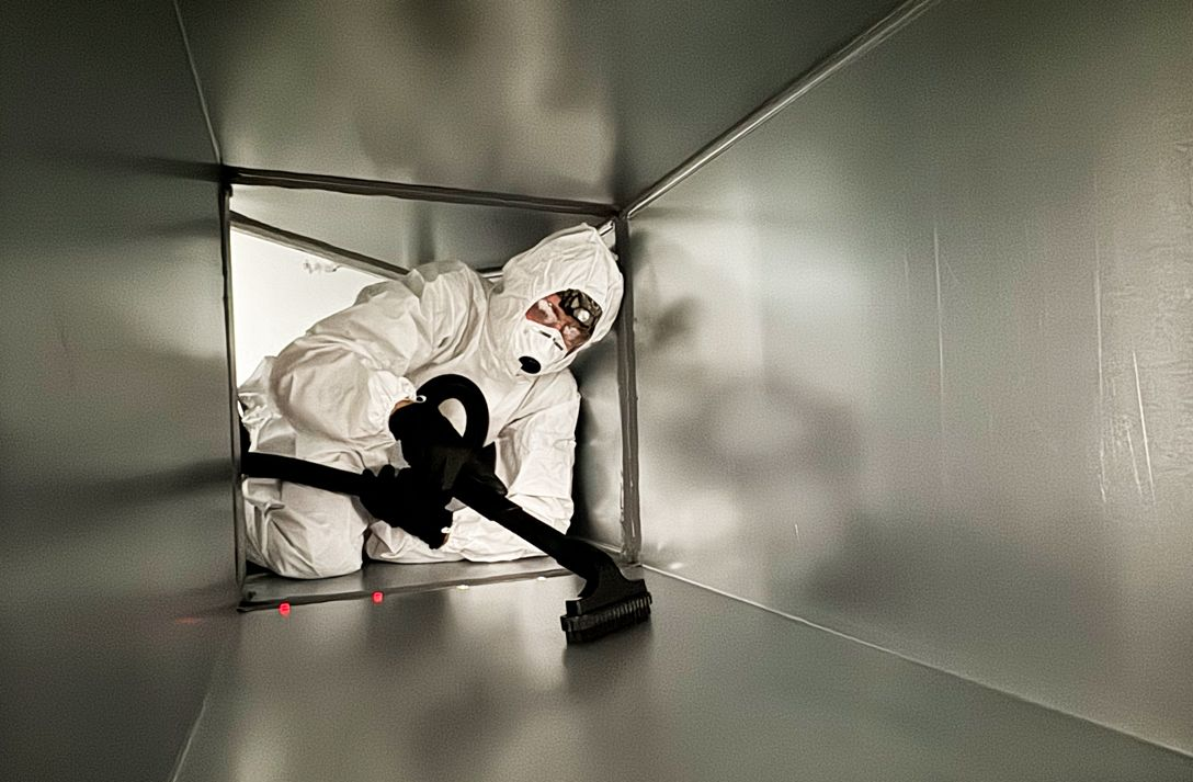 Person in white protective suit cleaning a metal ventilation duct with a black hose.
