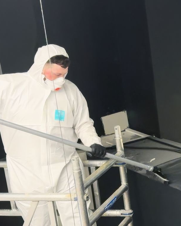 Person in white hazmat suit on scaffolding, cleaning, wearing a mask. Black wall in background.