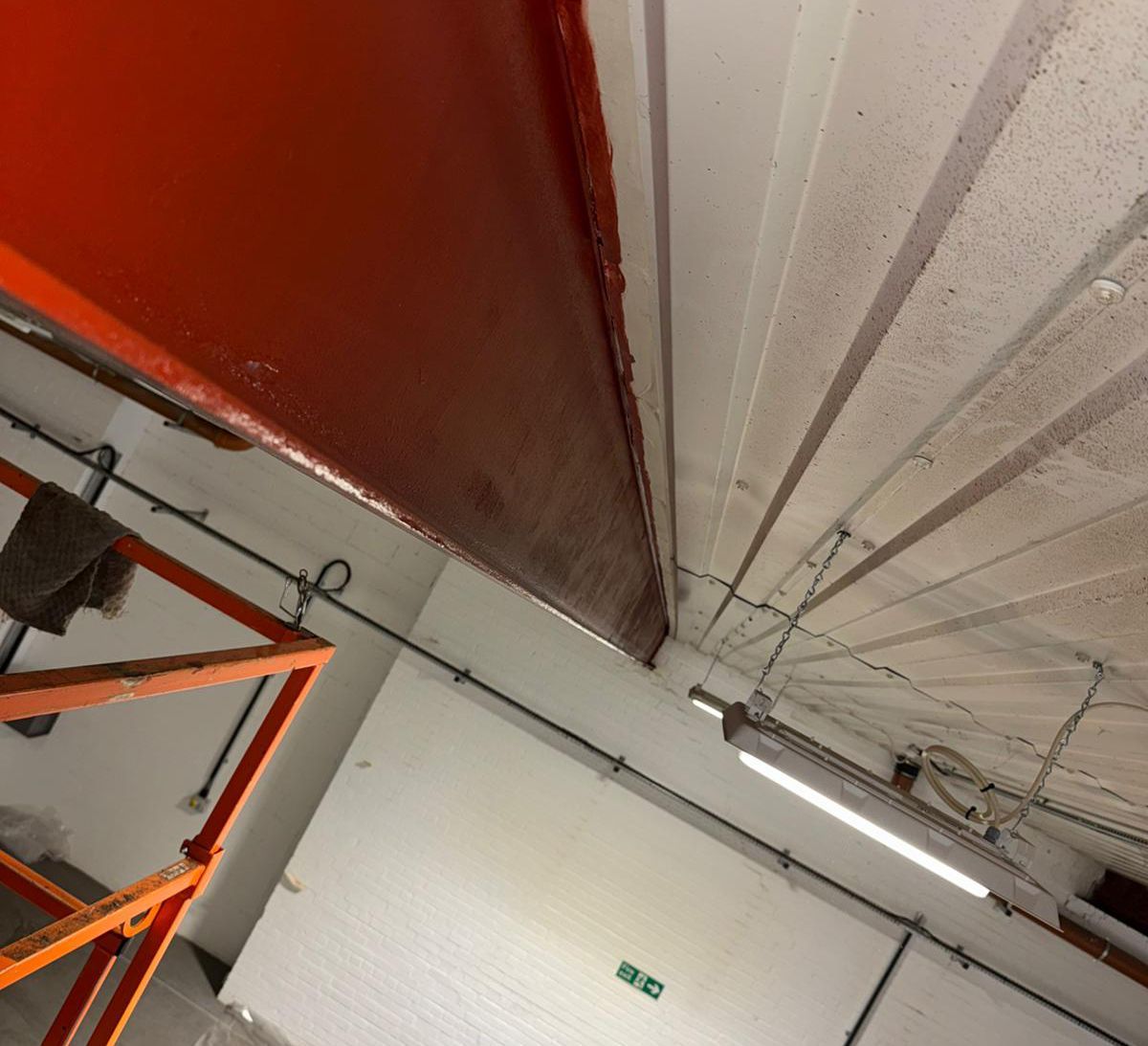 Orange painted metal beam in a white room. Electrical wiring and fluorescent lights are visible.