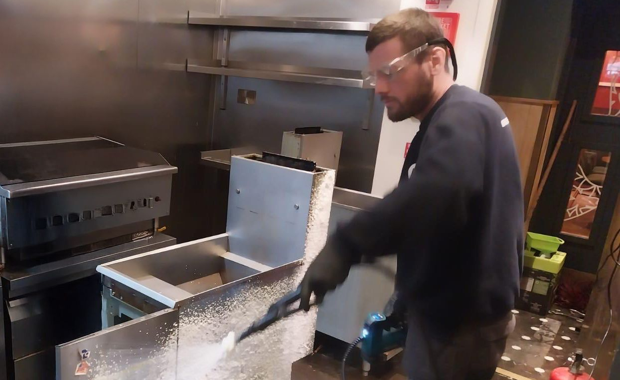 Person in a kitchen opening a stainless-steel grill or fryer hood beside another metal appliance.