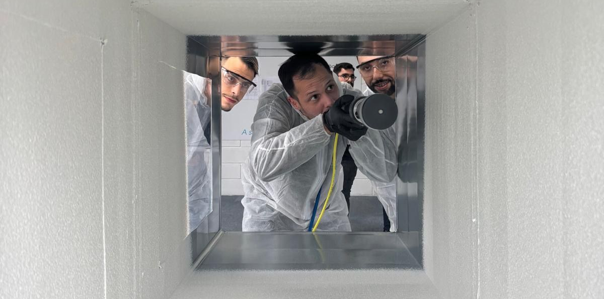 Inside a white rectangular space, three people in white suits and gloves work with a tool and wires on a metal surface.