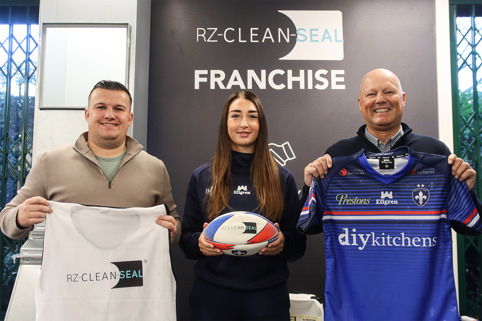 Three people holding sports jerseys and a rugby ball, in front of a franchise logo.