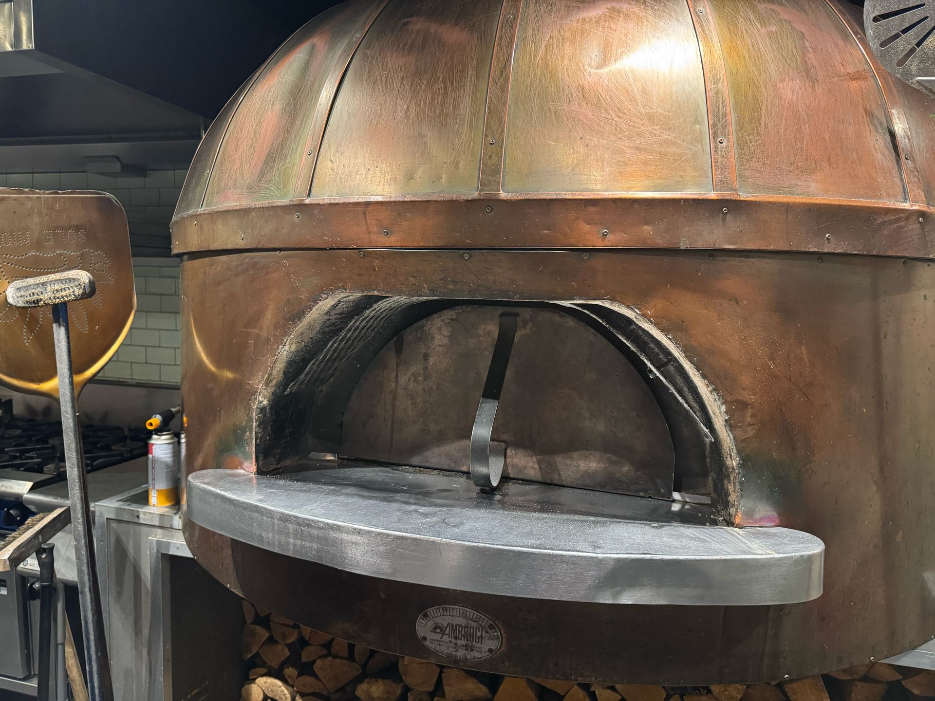 Copper pizza oven with open mouth, shovel, and wood pile in a kitchen setting.