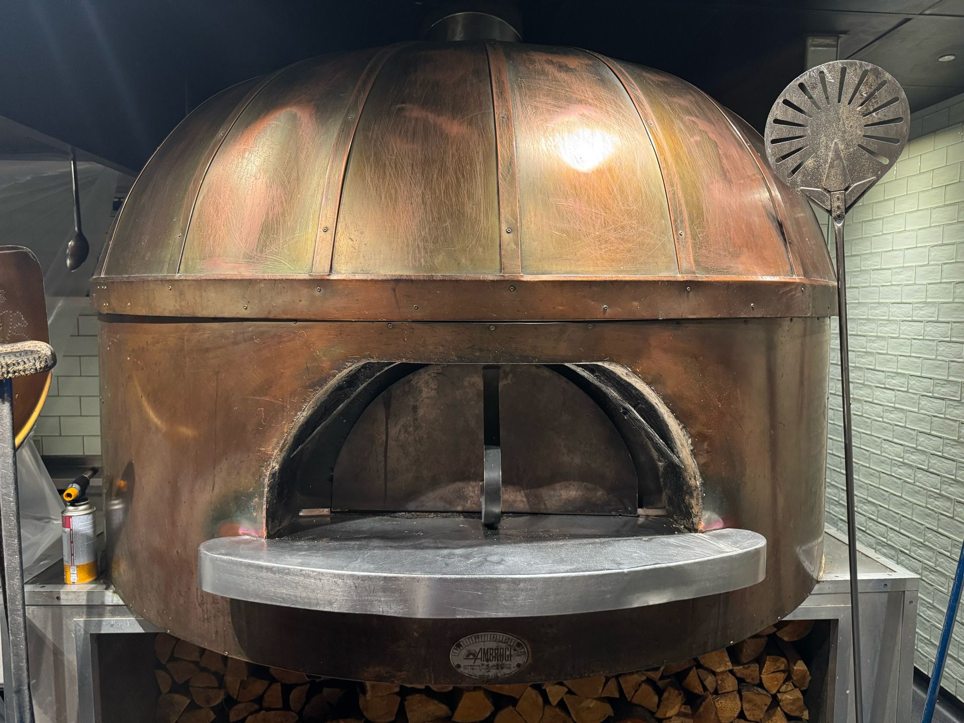 Copper-domed wood-fired pizza oven with open mouth, stone base, and pile of wood underneath.