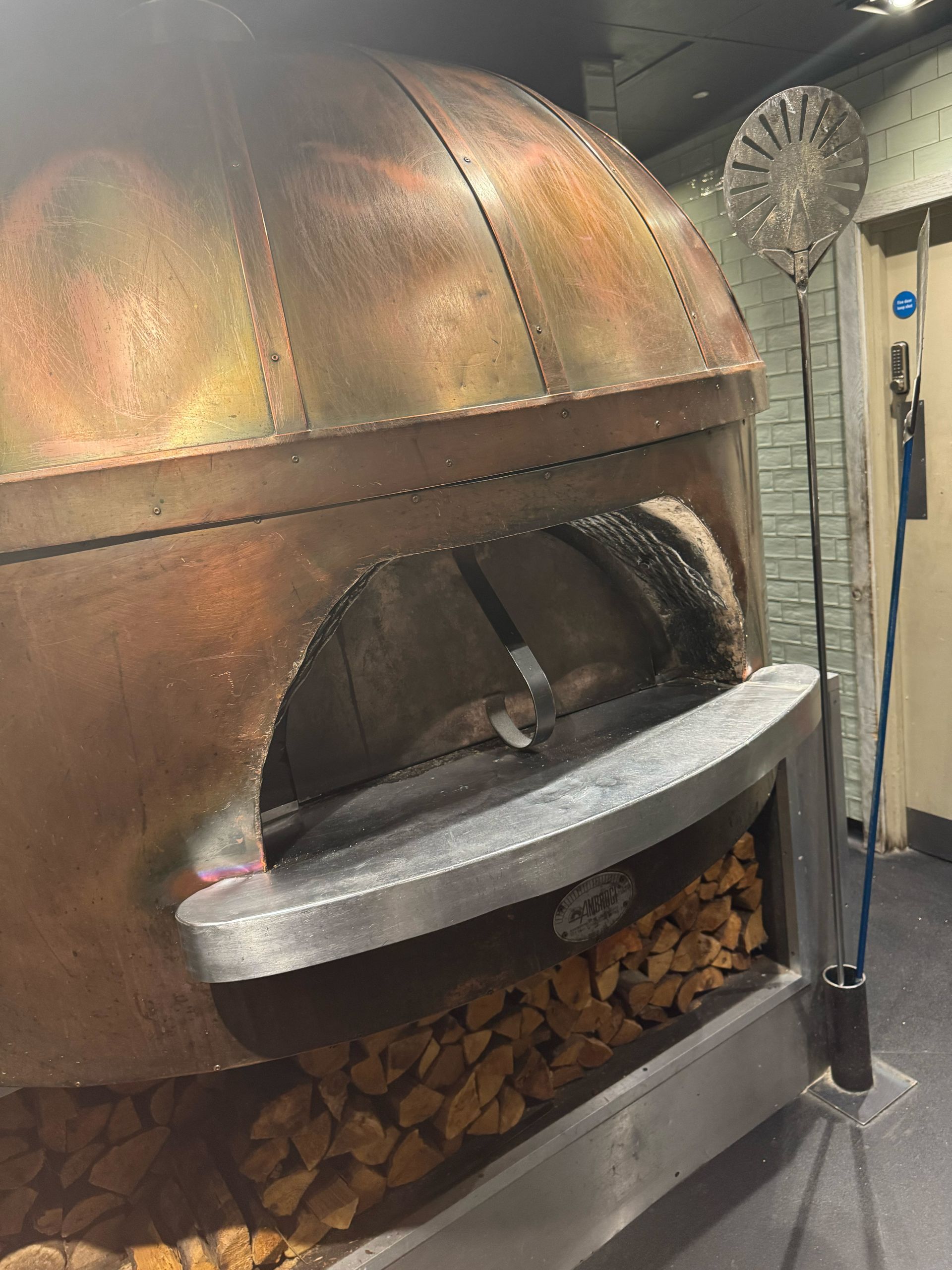 Copper-colored wood-fired pizza oven with a wide opening and a pile of wood underneath.
