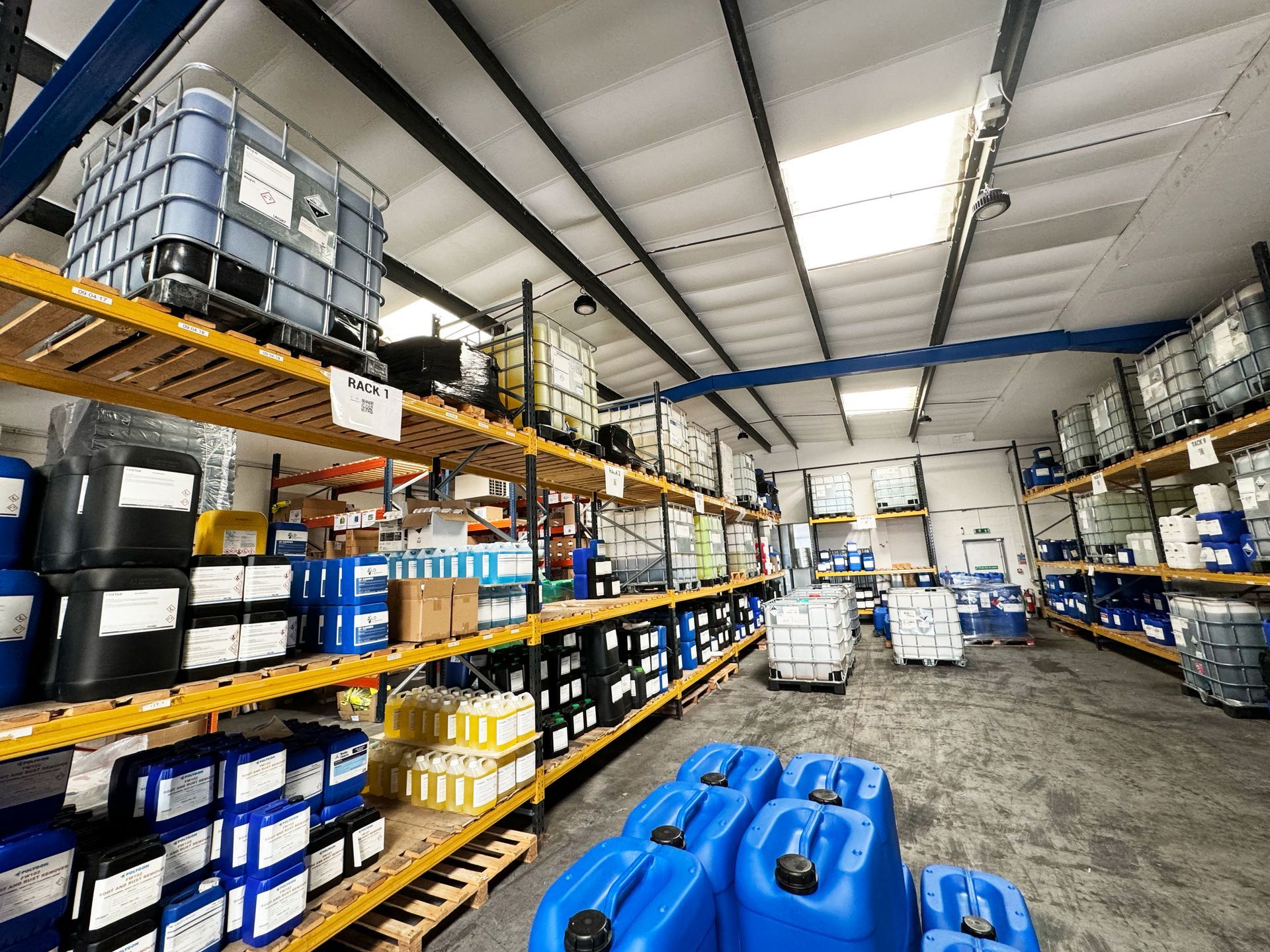 Industrial warehouse aisle with metal racks, stacked containers, and blue plastic tanks in the foreground