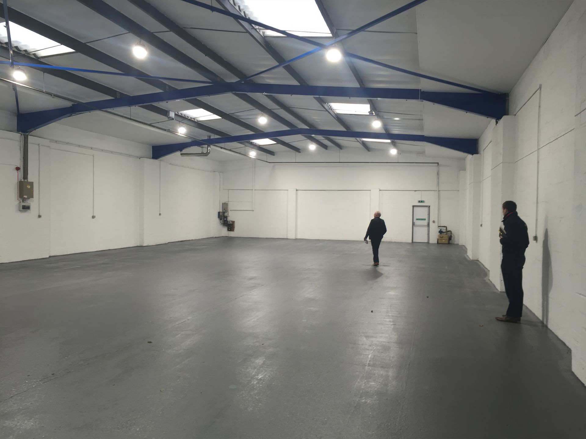 Large, empty warehouse interior with two people. White walls, grey floor, blue beams, and skylights.