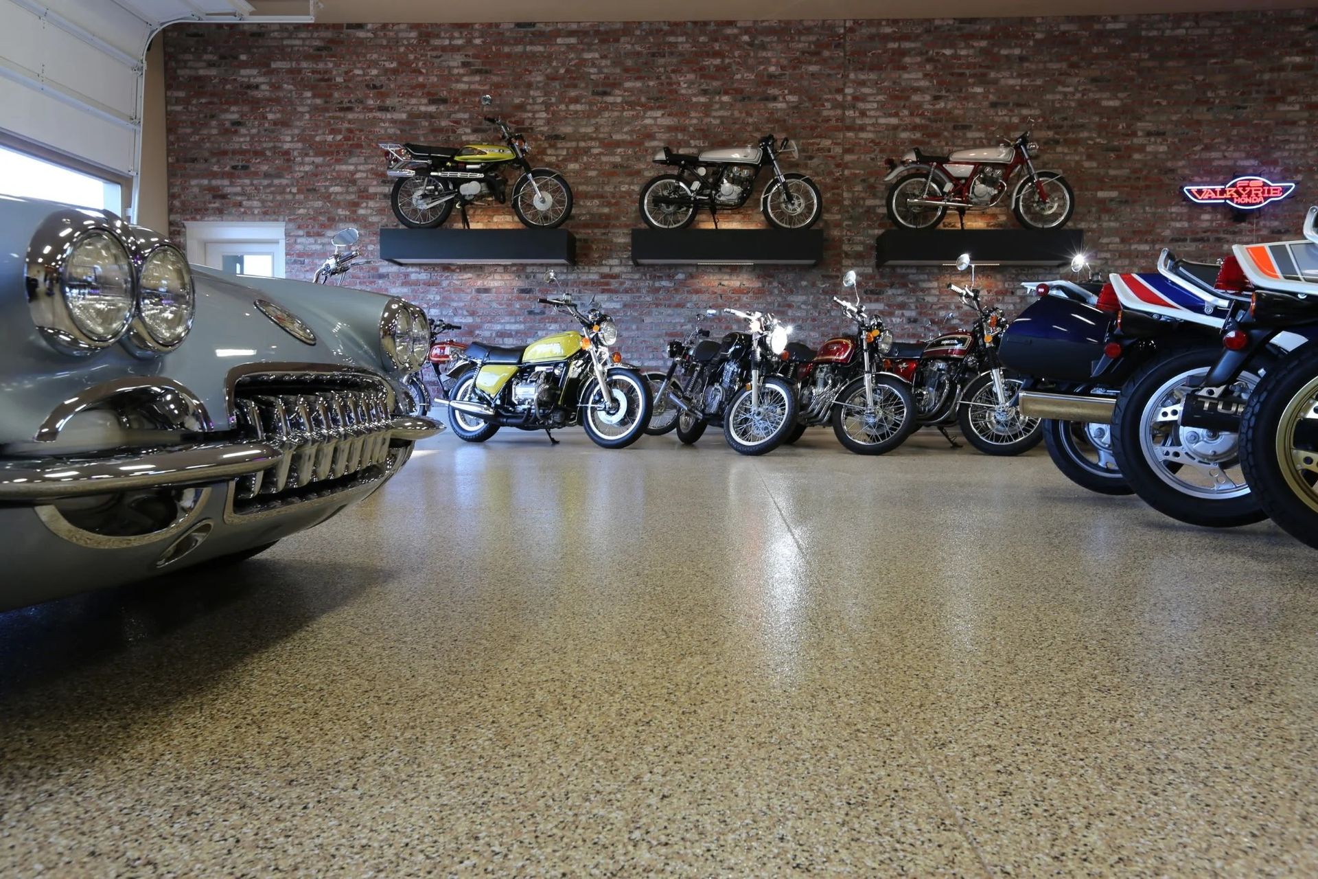 A showroom with classic motorcycles and a vintage car on a speckled floor. Brick wall in the background.