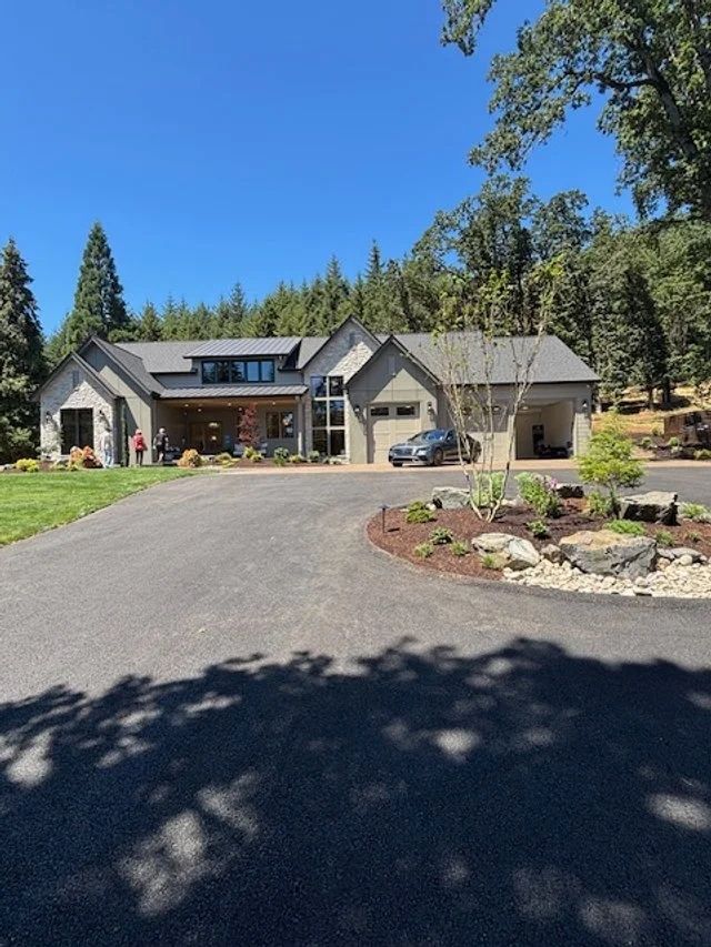 Modern gray house with black asphalt driveway and attached garage, in a wooded setting.