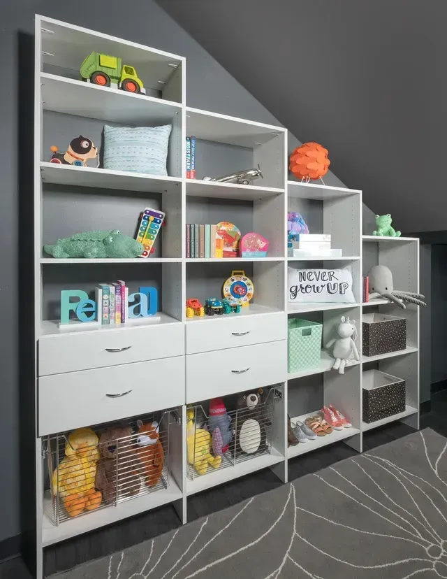 White storage shelves built into sloped wall, displaying toys and drawers, in a room with gray walls and carpet.