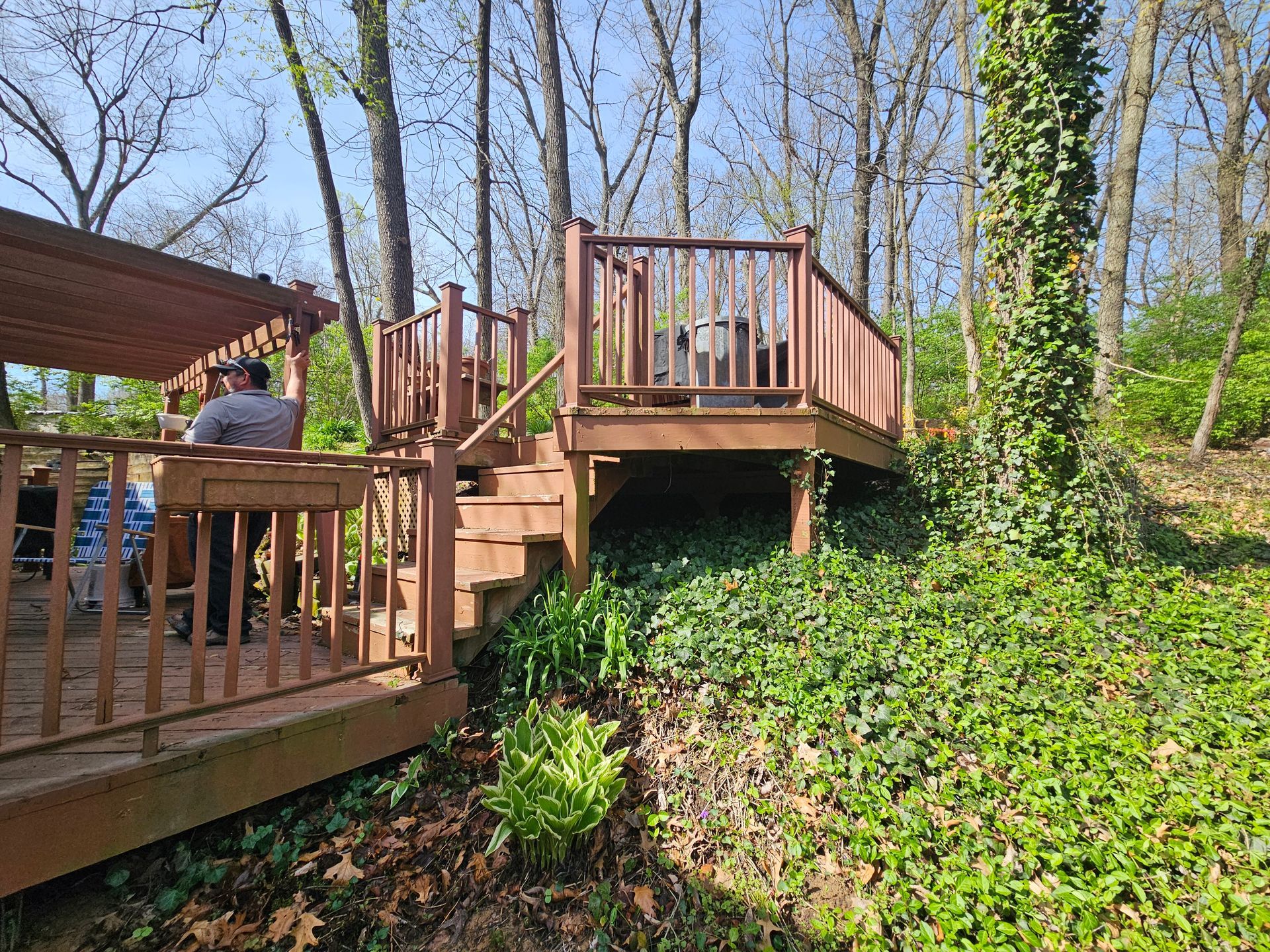 Wooden deck with stairs, surrounded by trees and greenery. A person is on the deck under a pergola.