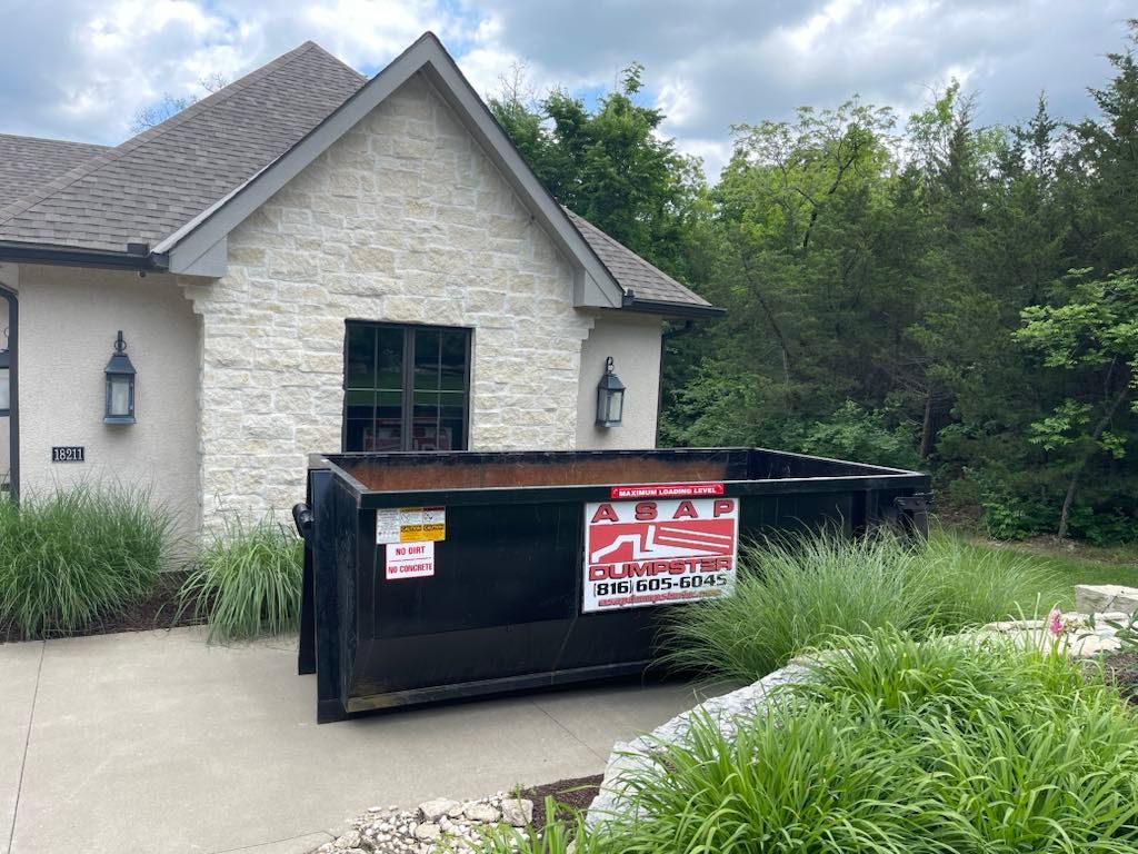 Black dumpster sits next to a stone building with a dark roof and green foliage.