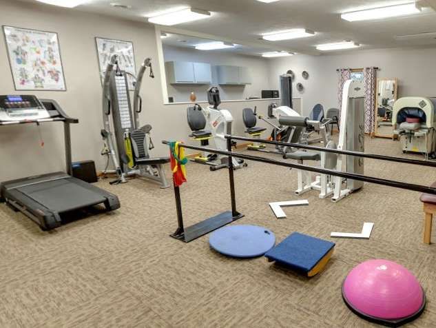 Physical therapy gym with exercise machines, parallel bars, balance pads, and a pink exercise ball.