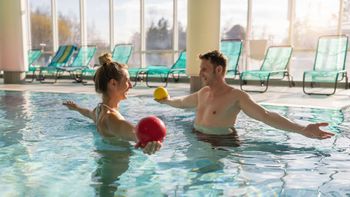 Two people in a pool playing with colorful balls, with lounge chairs in the background.