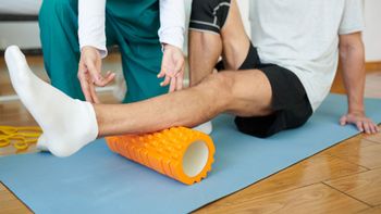 Two people doing a leg stretch with a foam roller on a yoga mat indoors
