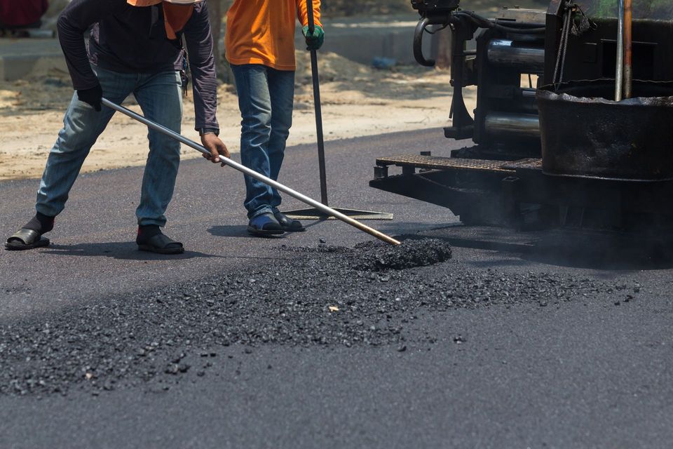 Workers shoveling hot asphalt beside a paving machine on a road repair site