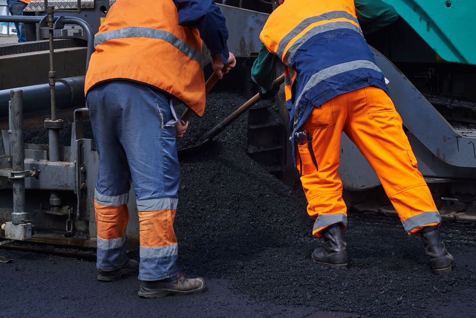Two road workers in orange reflective gear spreading asphalt on a road paver