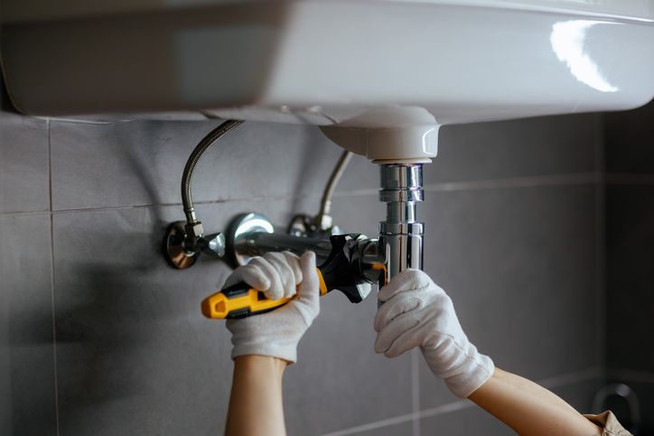 A plumber tightens a chrome drain pipe under a white sink with a wrench. A plumber tightens a chrome drain pipe under a white sink with a wrench.