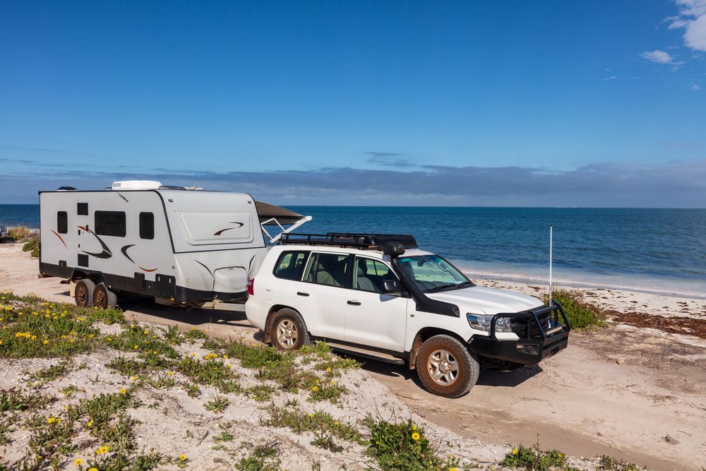 White SUV towing a grey caravan on a beach, blue sea and sky. — Caravan and Trailer Clinic Geelong in Breakwater, VIC