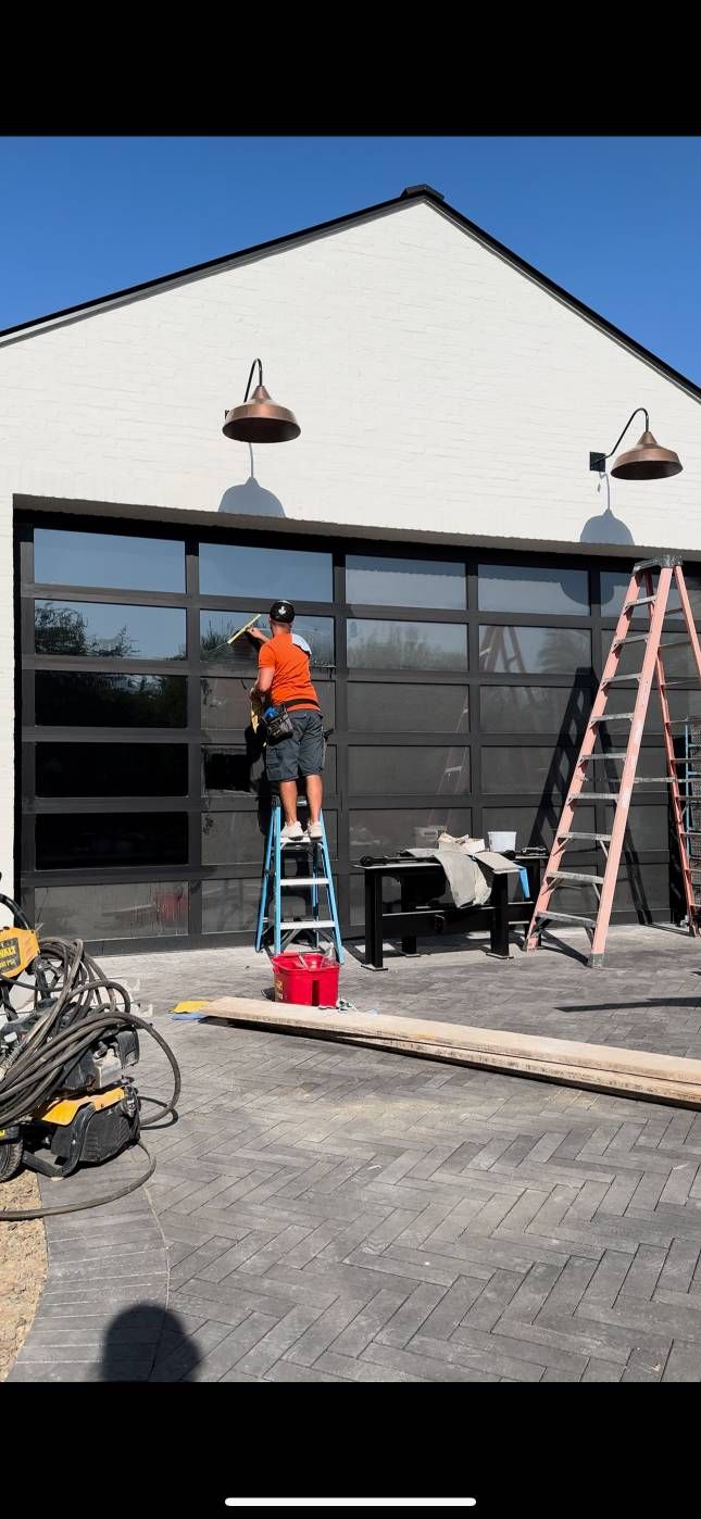 A worker on a ladder cleaning a glass garage door. Tools and materials are nearby. Blue sky.